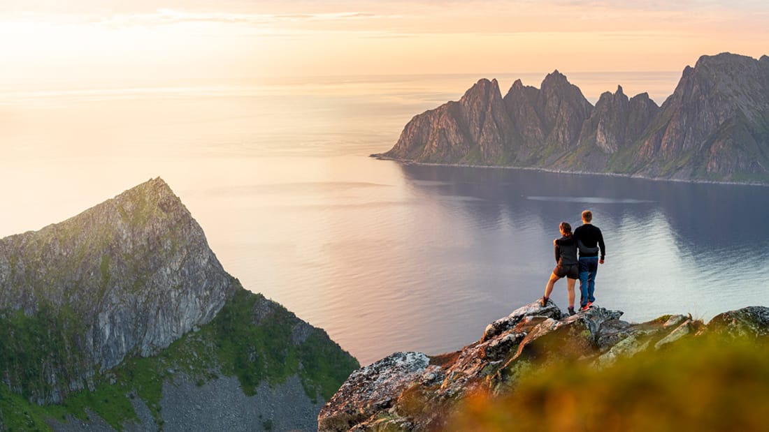 To personer står på toppen av et fjell og ser utover havet.