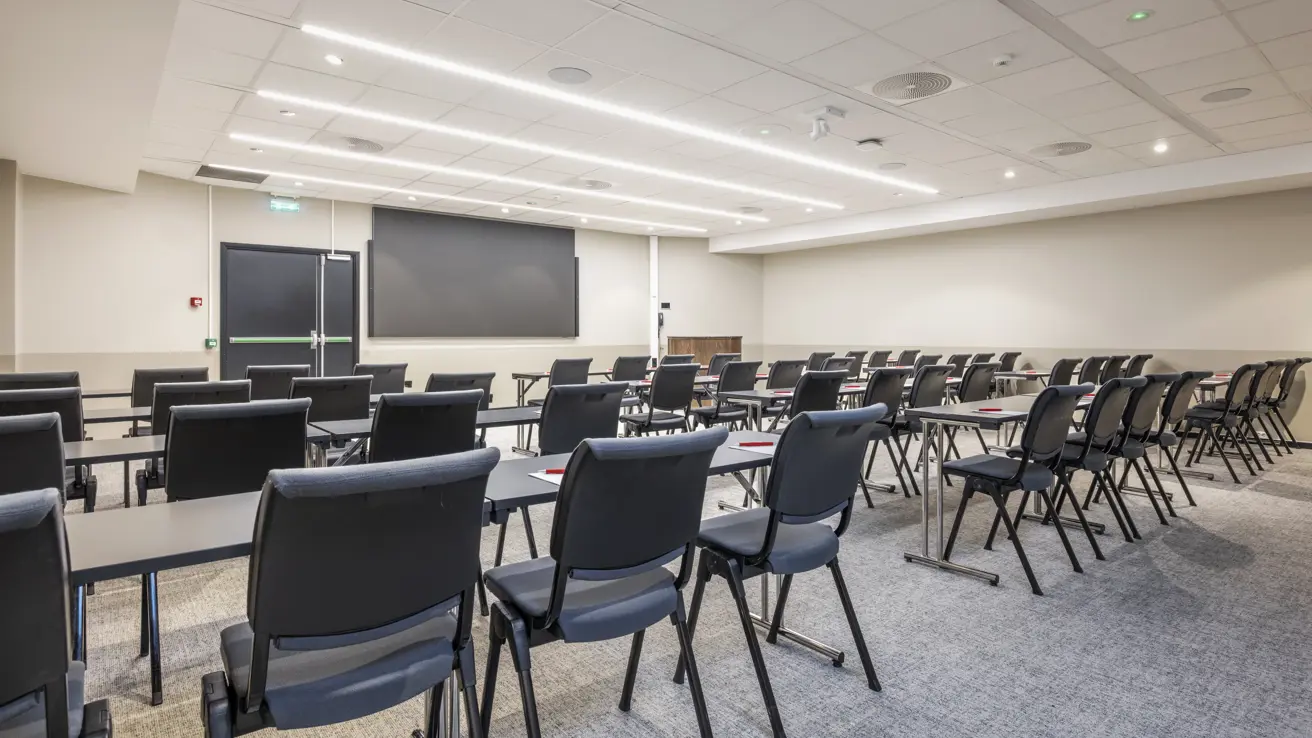 Konferanserom oppsett med stoler bak bord og en skjerm forrest i rommet.