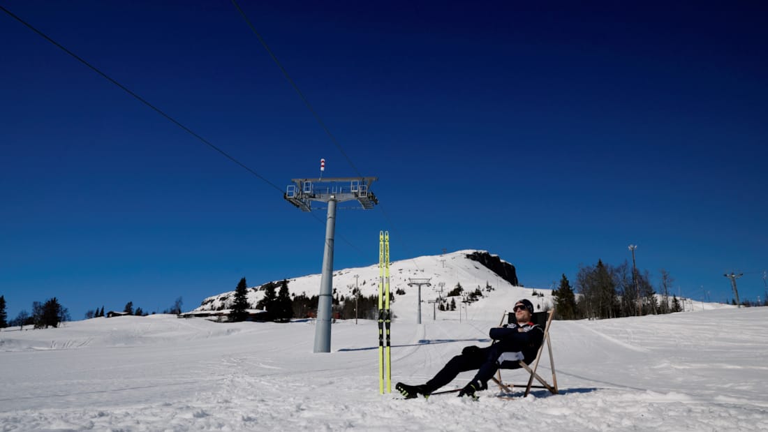 Mann som sitter i en stol i skibakken i Skeikampen alpinsenter. Snødekt fjell og blå himmel i bakgrunnen. 