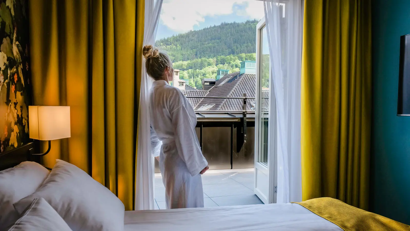Kvinne i morgenkåpe som står i døråpningen til balkong på et hotellrom. Gule, store gardiner på hver side av døren.