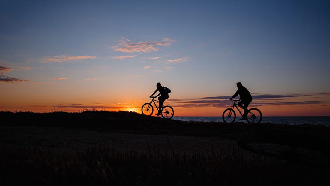 Silhuett av to syklister sykler på en voll ved sjøen, i solnedgang.