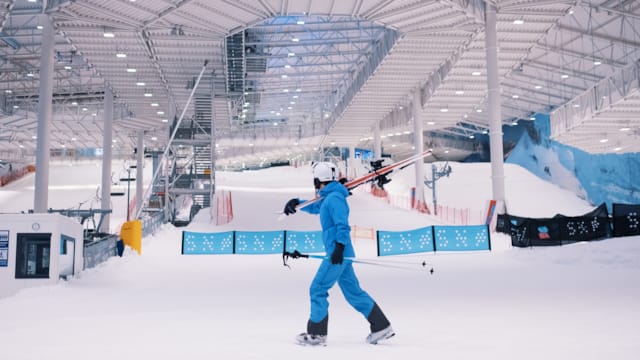Mann iført blå skidrakt bærer på et par med ski i Thon Snø innendørs skiarena