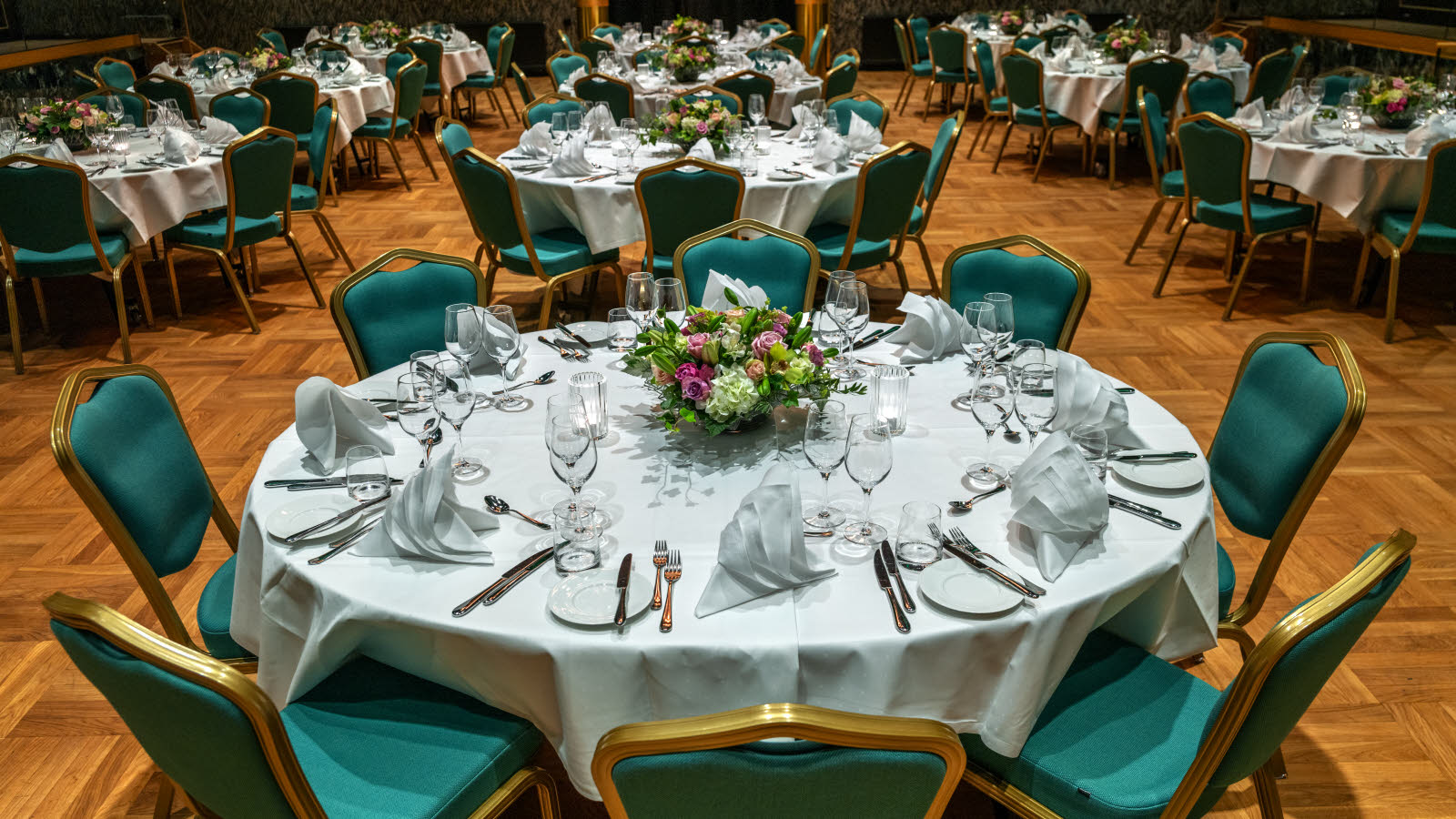 Flagstadsalen selskapsoppsett med runde bord og grønne stoler. Bord trukket med hvit duk og oppsett til 8 personer med blomsterbukett midt på bordet.