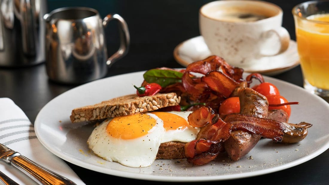 Nærbilde av tallerken med brød, tomater, egg og bacon. Kaffe og glass med appelsinjuice i bakgrunnen.