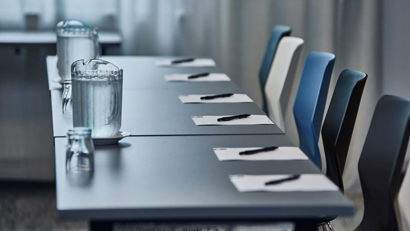 Nærbilde av skrivebord og stoler. Bordet er dekket med skrivesaker, vannglass og vannmugge.