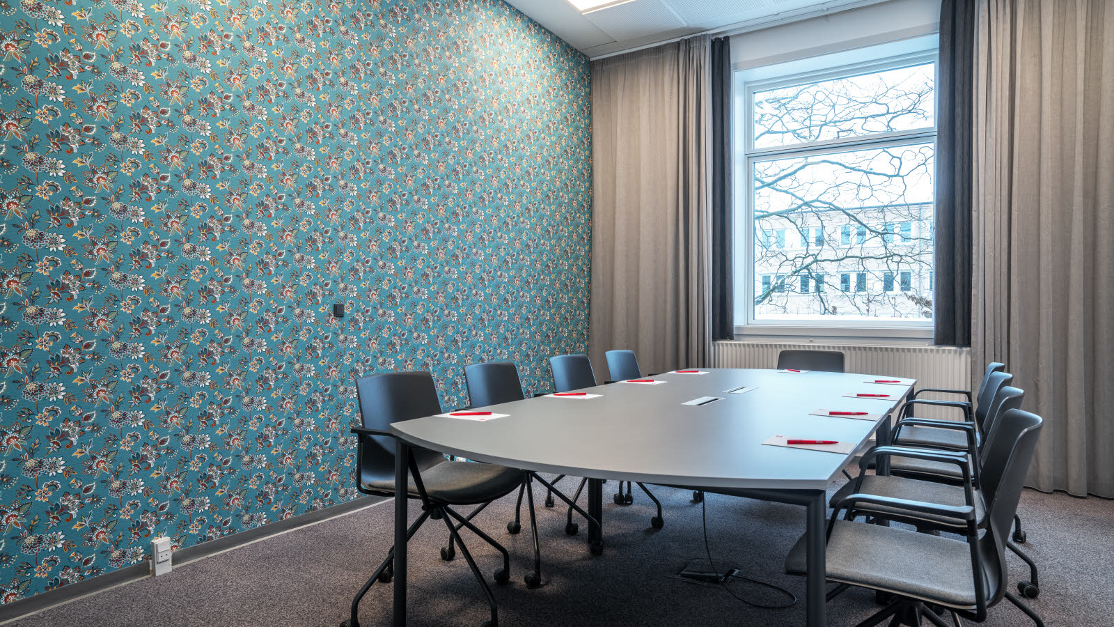 Bord med 9 kontorstoler på hjul rundt. Skrivesaker på bordet. Vindu og gardiner. Blå tapet med blomstermønster i rødt, hvit grønt og gult.