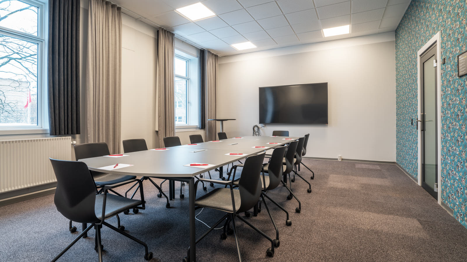 Møterom 8 med to vinduer på langvegg, TV-skjerm på kortvegg, styrebordsoppsett med tolv kontorstoler på hjul rundt. Tapetvegg og glassdør til høyre.