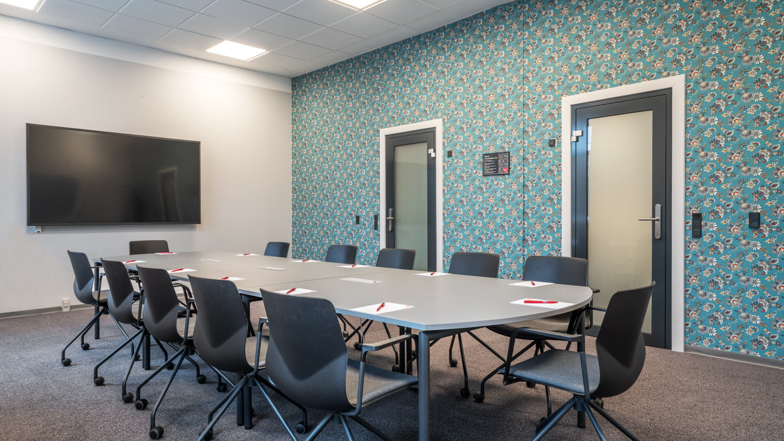 Styrebord med 12 sorte kontorstoler på hjul rundt. Skrivesaker på bordet. TV-skjerm på kortvegg. Langvegg med blå tapet med blomster på. To glassdører på langvegg. Teppebelagt gulv.
