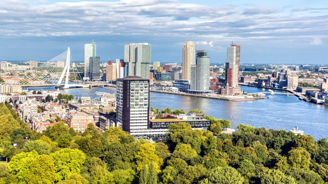 Oversiktsbilde over Rotterdam. Skog i forgrunnen, elv, skyskrapere og bro i bakgrunnen. Blå himmel med gråhvite skyer.