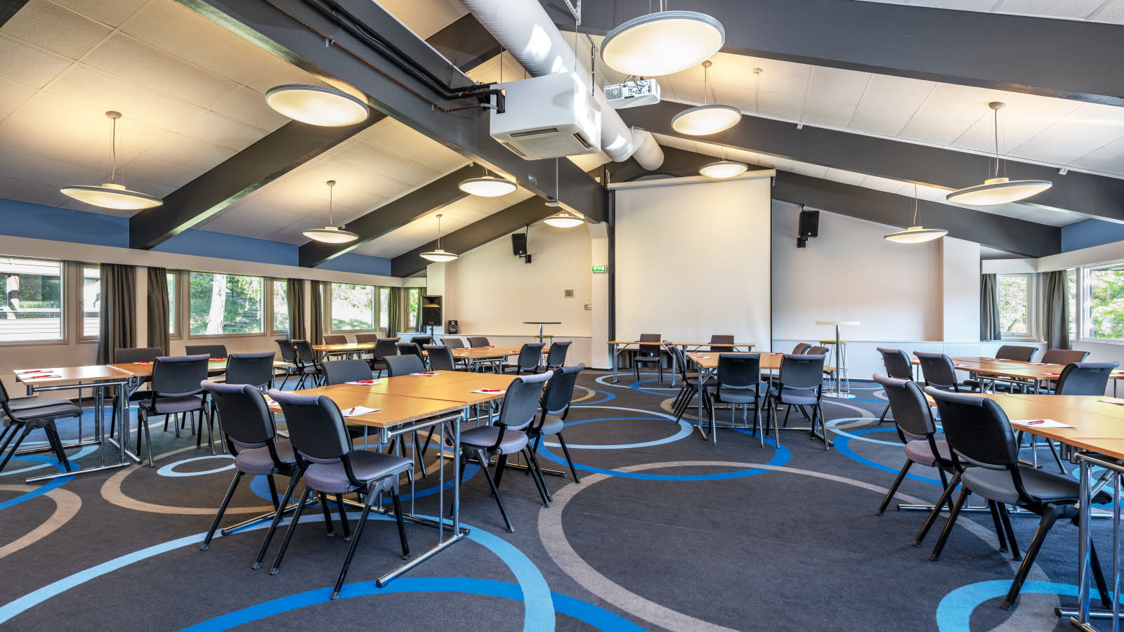 Konferansesal med gruppe bord, store taklamper, prosjektor og lerret og teppebelagt gulv.