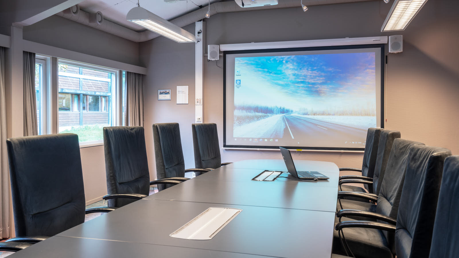Møterom i styrebordsoppsett, lerret i front og vinduer på langside.