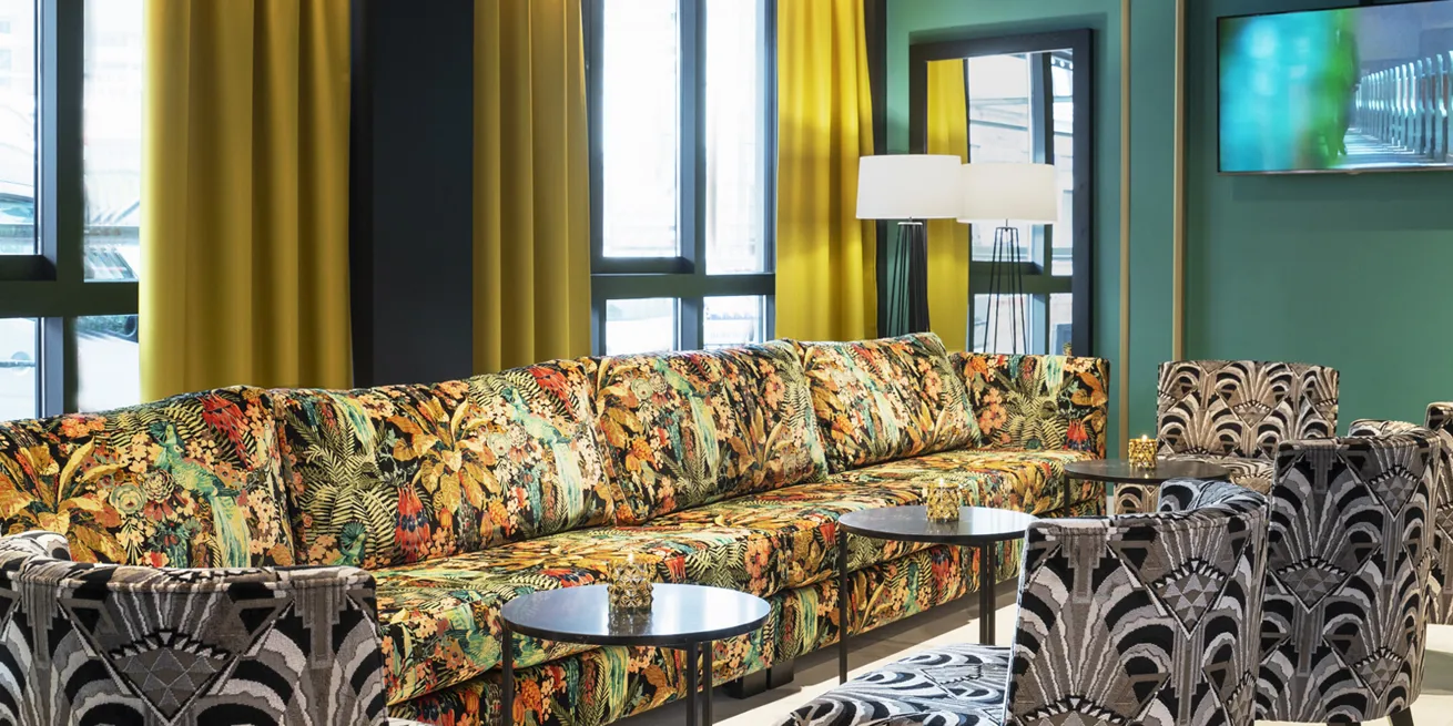 Sofaomr&aring;de i en hotellobby med blomstrete trekk p&aring; sofaen, gr&oslash;nne vegger og gule lange gardiner.