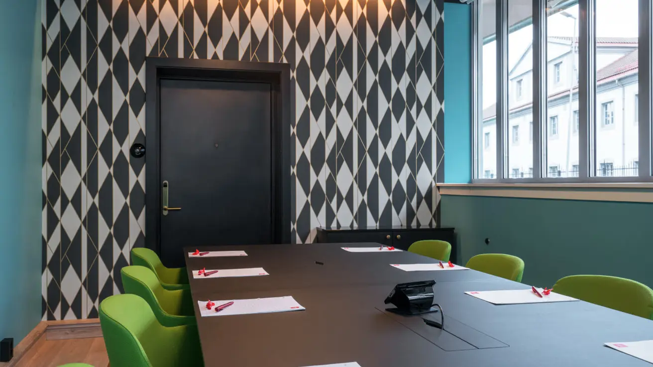 Møterom med skrivebordsoppsett. Sort bord med åtte, grønne stoler rundt. Skrivesaker og drops ligger dandert på bordet ved hver sitteplass. Kortvegg med mønstrete tapet i sort-hvitt. Dør på kortvegg. Projektor i taket, vinduer på langside. Parkettgulv.