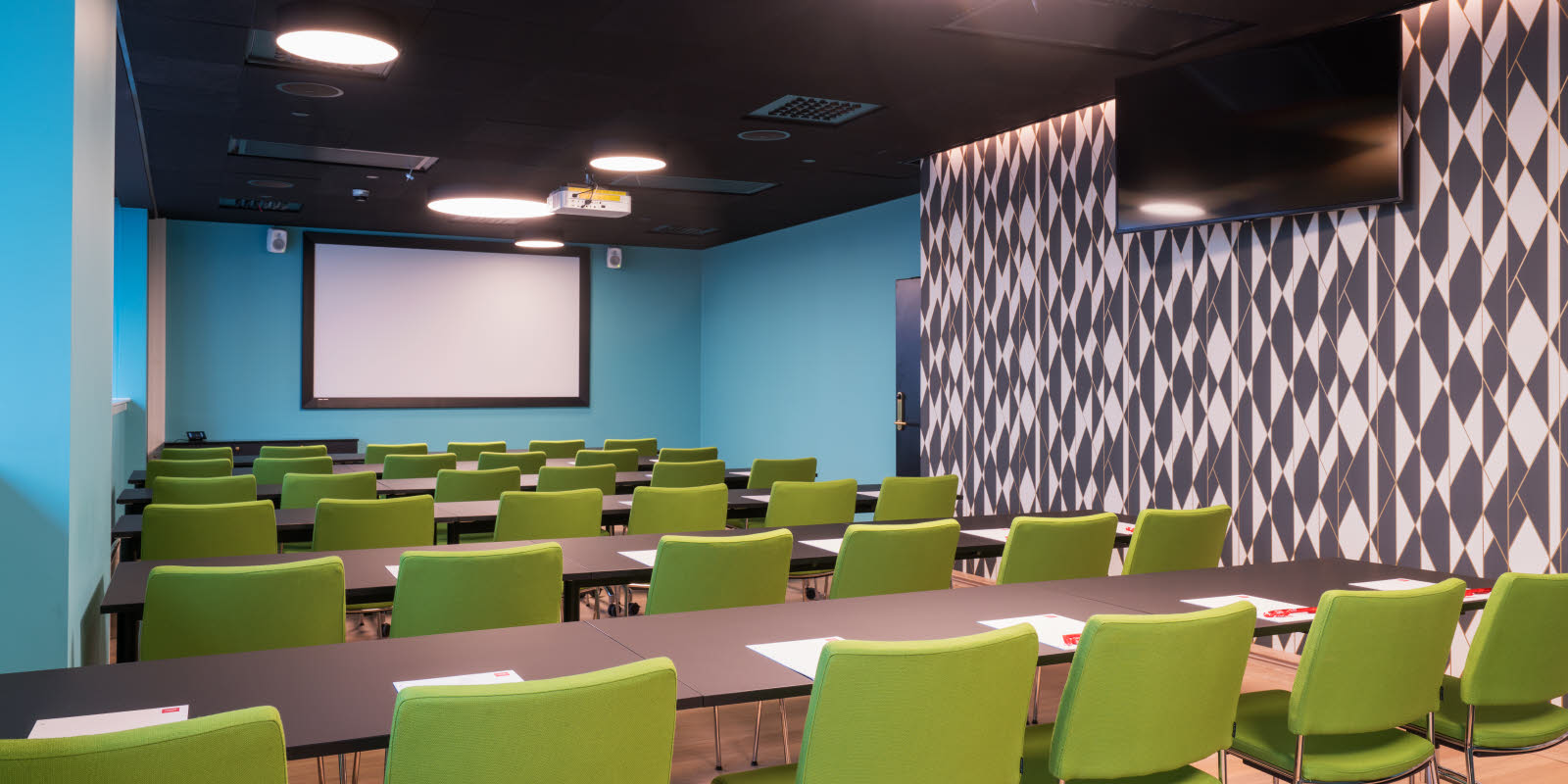 Konferanserom med klasseromsoppsett, sort og hvit mønstrete tapet på langsiden, turkise vegger i front og langside. Lerret i front, skjerm på langside, vinduer på langside.