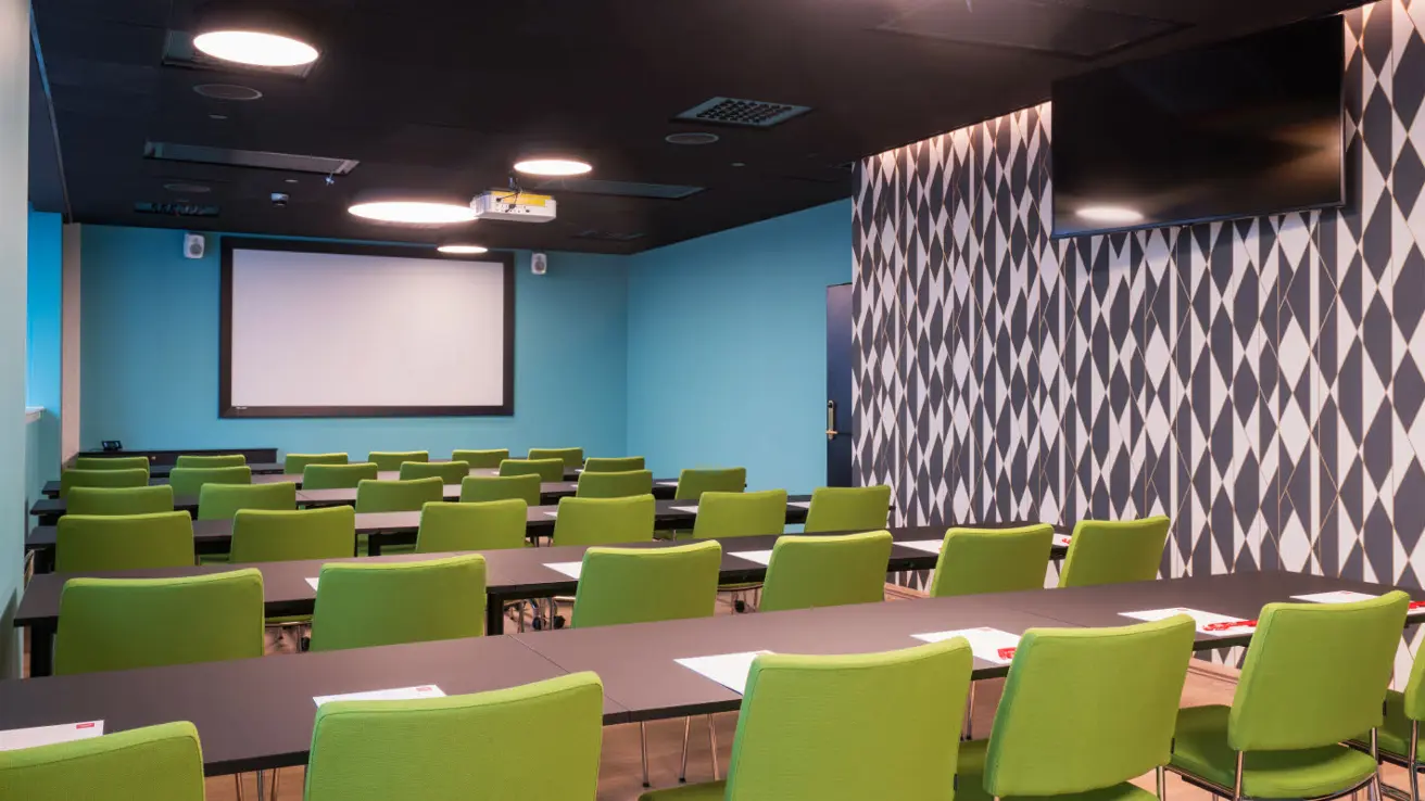 Konferanserom med klasseromsoppsett, sort og hvit mønstrete tapet på langsiden, turkise vegger i front og langside. Lerret i front, skjerm på langside, vinduer på langside.