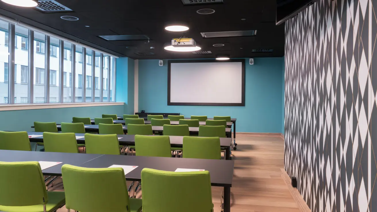 Møterom i klasseromsoppsett med vinduer langs hele langsiden, mønstrete tapet på motsatt siden, lerret i front.