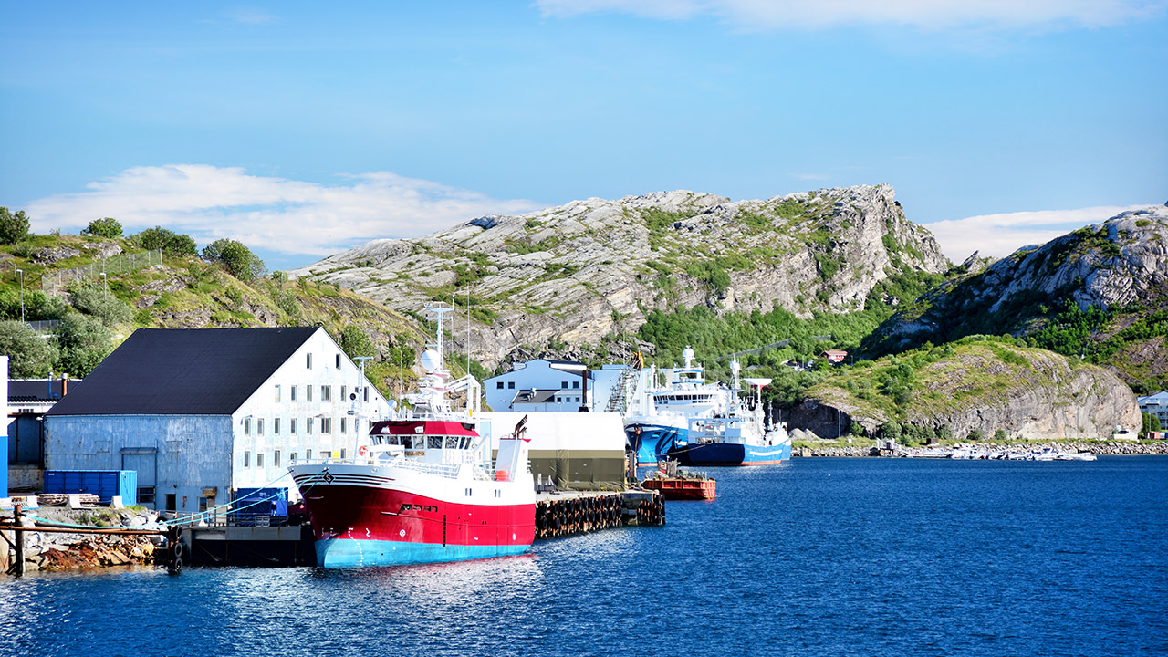 Båt ligger til kai foran bebyggelse. Fjell i bakgrunnen.