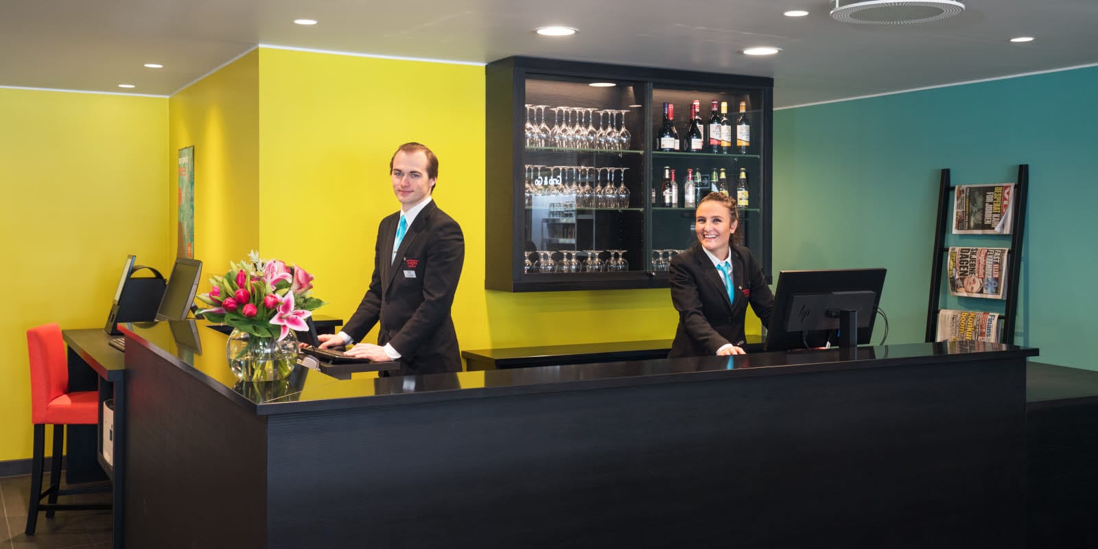 Resepsjon med gule og turkise vegger med to smilende resepsjonister på Thon Hotel Gardermoen