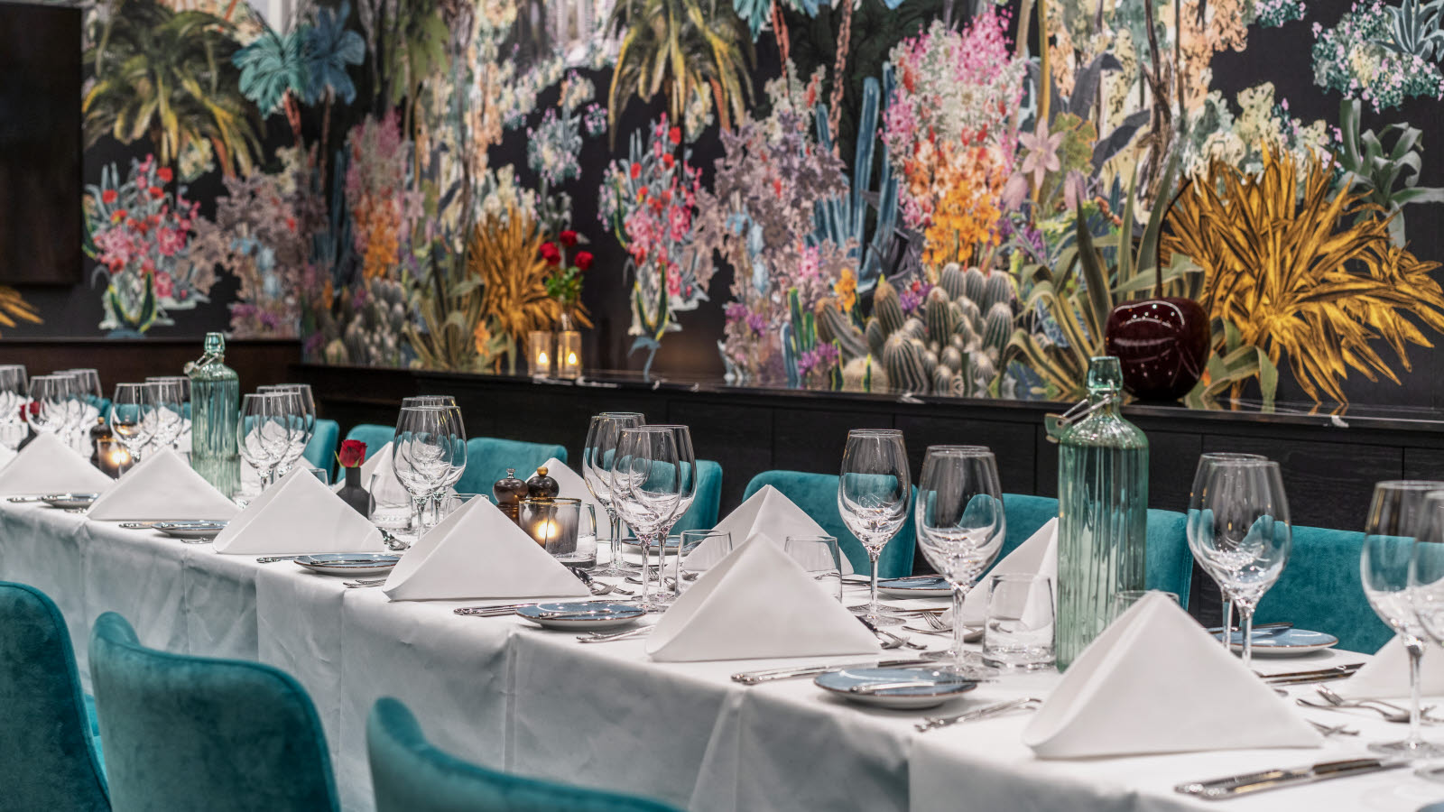 Chambre Sepaée med langbord dekket med hvit duk, hvise servietter, bestikk og glass. Tapet med plamer og blomster i ulike farger. Turkise stoler.