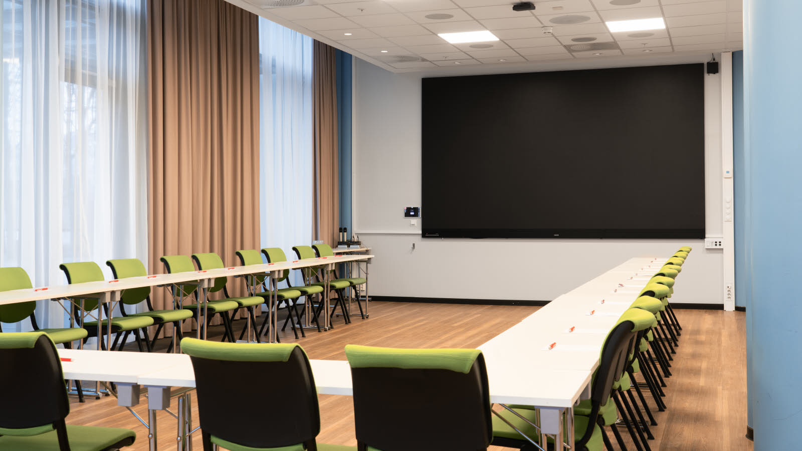 Møterom i hesteskooppsette med skrivebord og stoler. Skjerm i front. Vinduer på den ene langsiden.