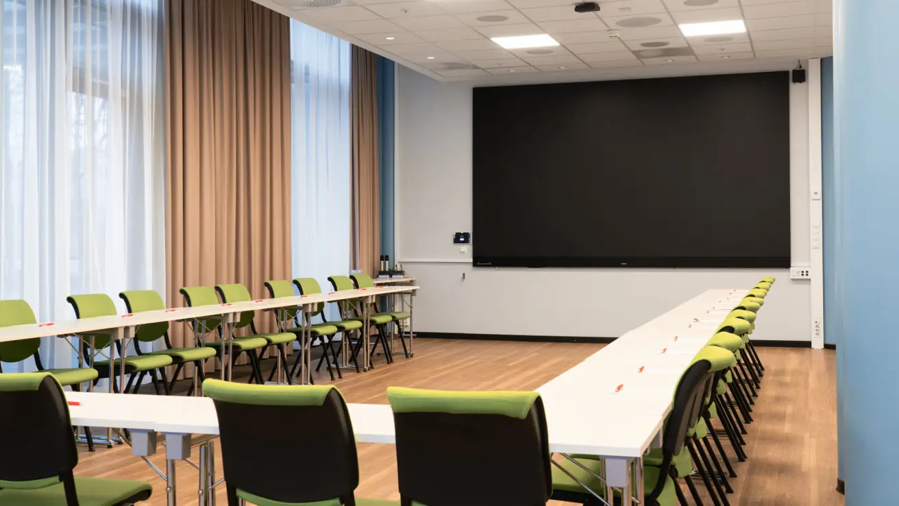 Møterom i hesteskooppsette med skrivebord og stoler. Skjerm i front. Vinduer på den ene langsiden.