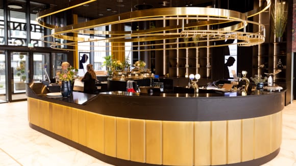 To resepsjonister i den ovalformet resepsjon på Thon Hotel Svolvær, med bar i andre enden. I bakgrunnen ses sitteplasser og kaffemaskin.