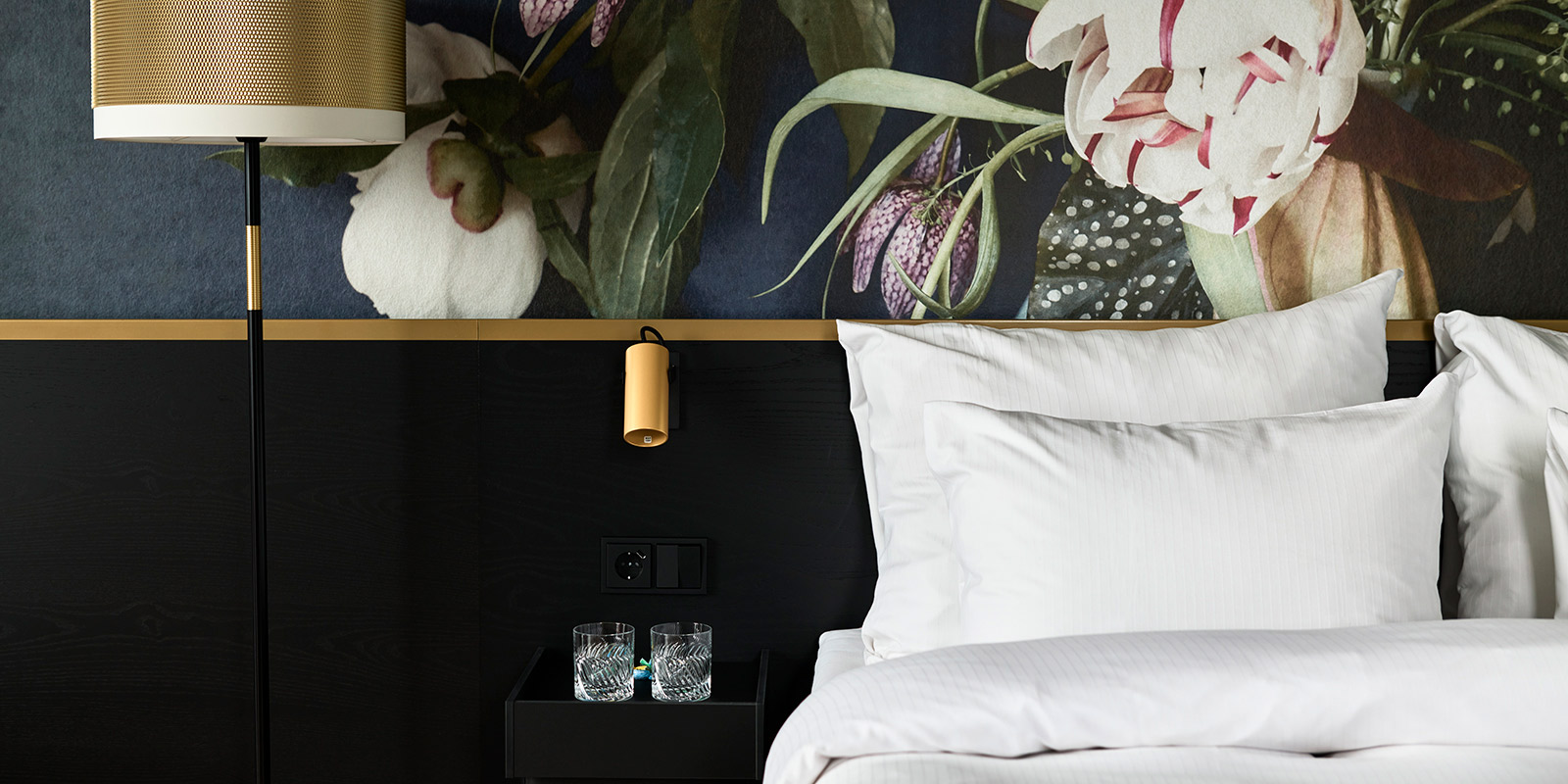 Nærbilde av oppredd seng med nattbord og lampe til venstre for sengen. Blomstrete tapet bak sengen.