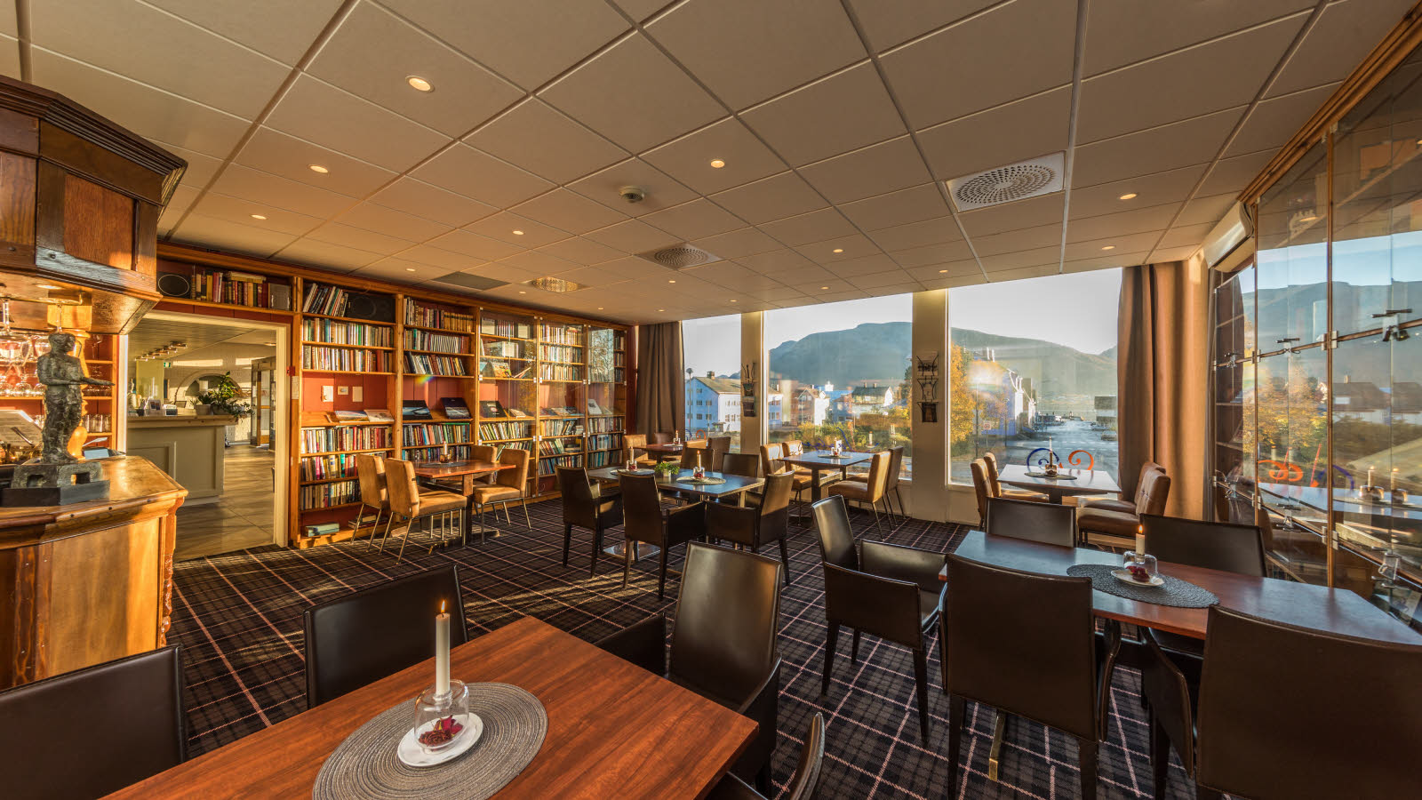 Hotellets bibliotek med flere bord og sitteplasser, teppebelagt gulv og store vindusflater.