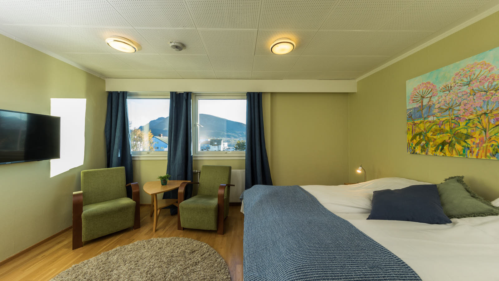 Standard Room Double med dobbeltseng, to lenestoler med bord mellom seg, TV, parkettbelagt gulv og to vinduer.