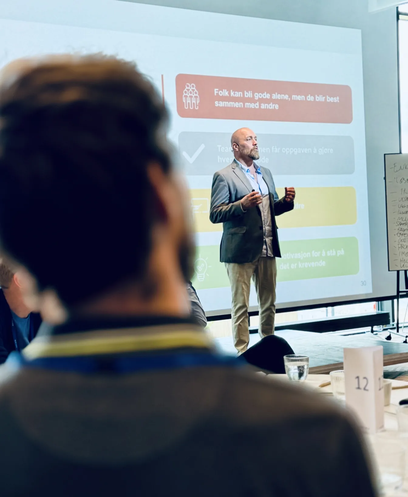 En mann i dress og skjorte st&aring;r foran en stor skjerm og holder et foredrag til en gruppe mennesker som sitter og h&oslash;rer p&aring;