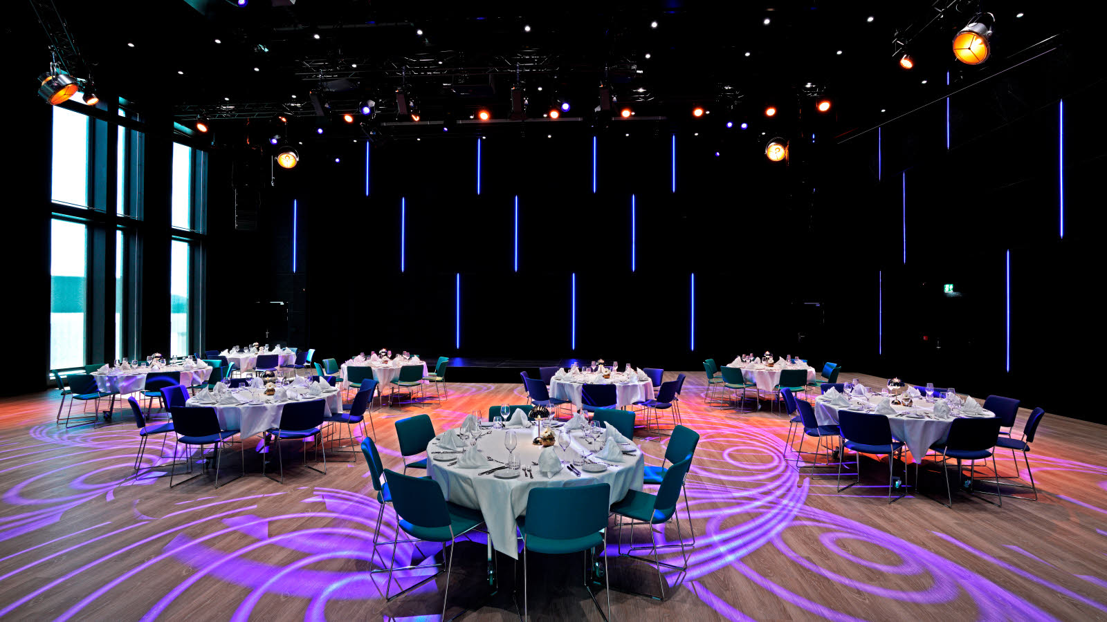 Konferansesal med bankettoppsett. Runde bord dekket med hvite duker, servise, bestikk, glass og servietter. På gulvet ses lilla mønster fra lyskasterne i taket. Store vinduer på den ene siden.