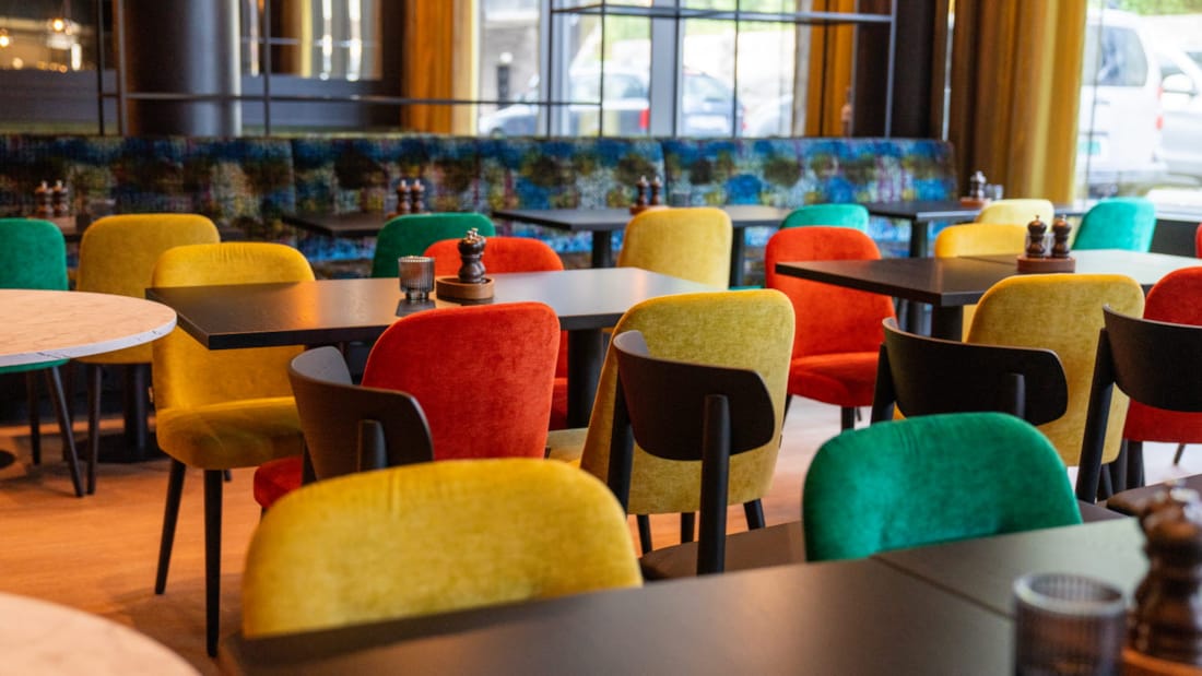 Restauranten hos Thon Hotel Snø.  Gule, Grønne og røde stoler rundt tilhørende, mørke bord. På bordene står telysholder og salt- og pepperkvern. I bakgrunnen sees en sofa i blågrønne  toner, og store vindusflater.