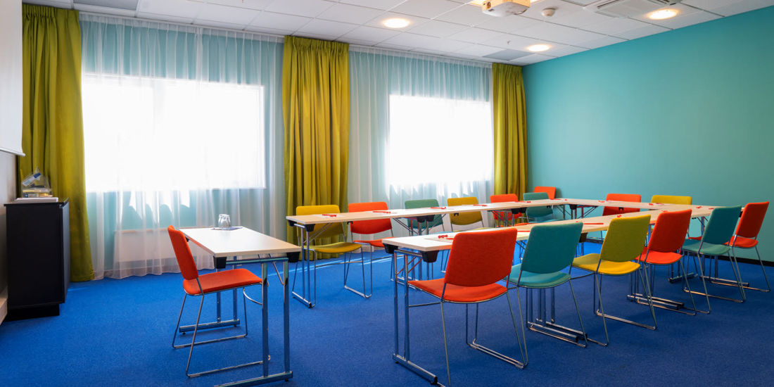Møterom øst 5 med blått teppe på gulvet, turkise vegger, sennepsgule gardiner, lerrett, prosjektor og fargerike stoler i hesteskooppsett på Thon Hotel Triaden i Lørenskog