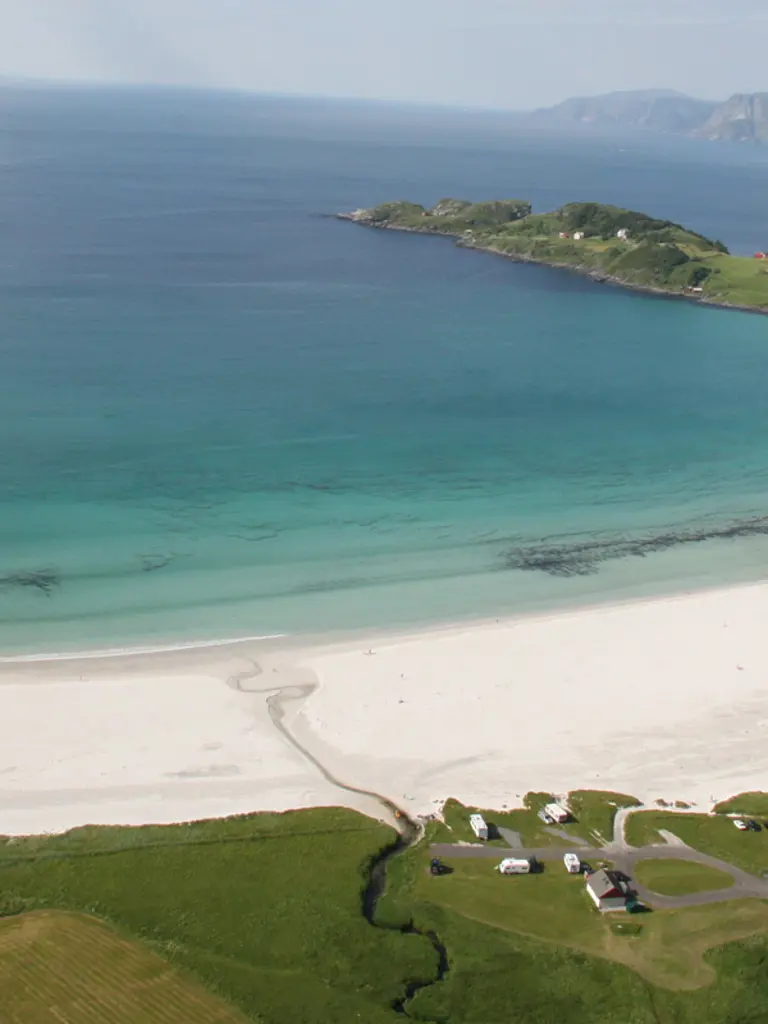 Bukt med hvit sandstrand og krystallblått hav, omkranset av grønt terreng, sett ovenfra.