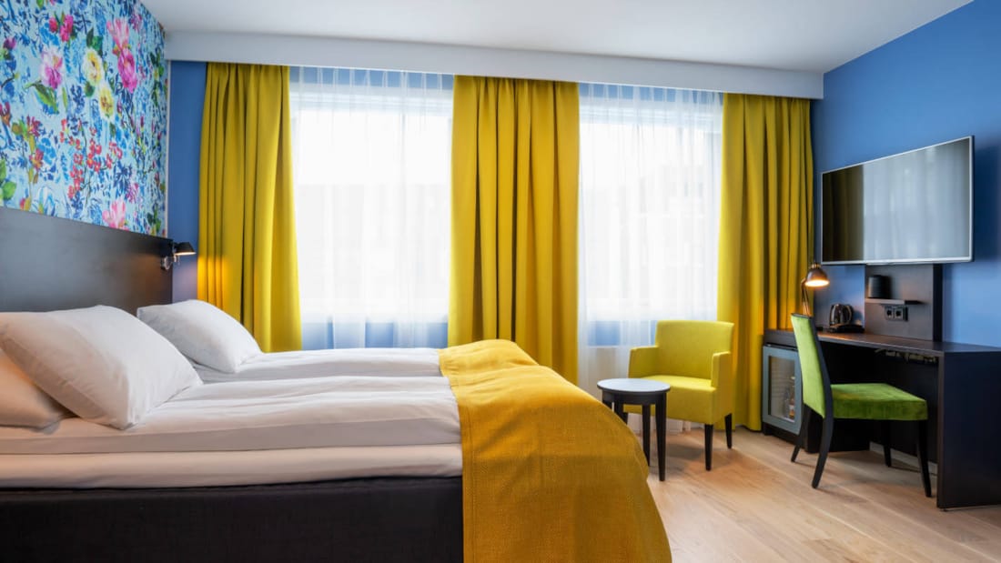 Rom med dobbeltseng, to store vinduer, TV, skrivebord, stol, blå tapet med blomster i ulike farger host Thon Hotel Moldefjord.