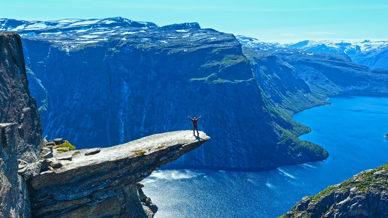 En person st&aring;r ytterst p&aring; Trolltunga i str&aring;lende sol