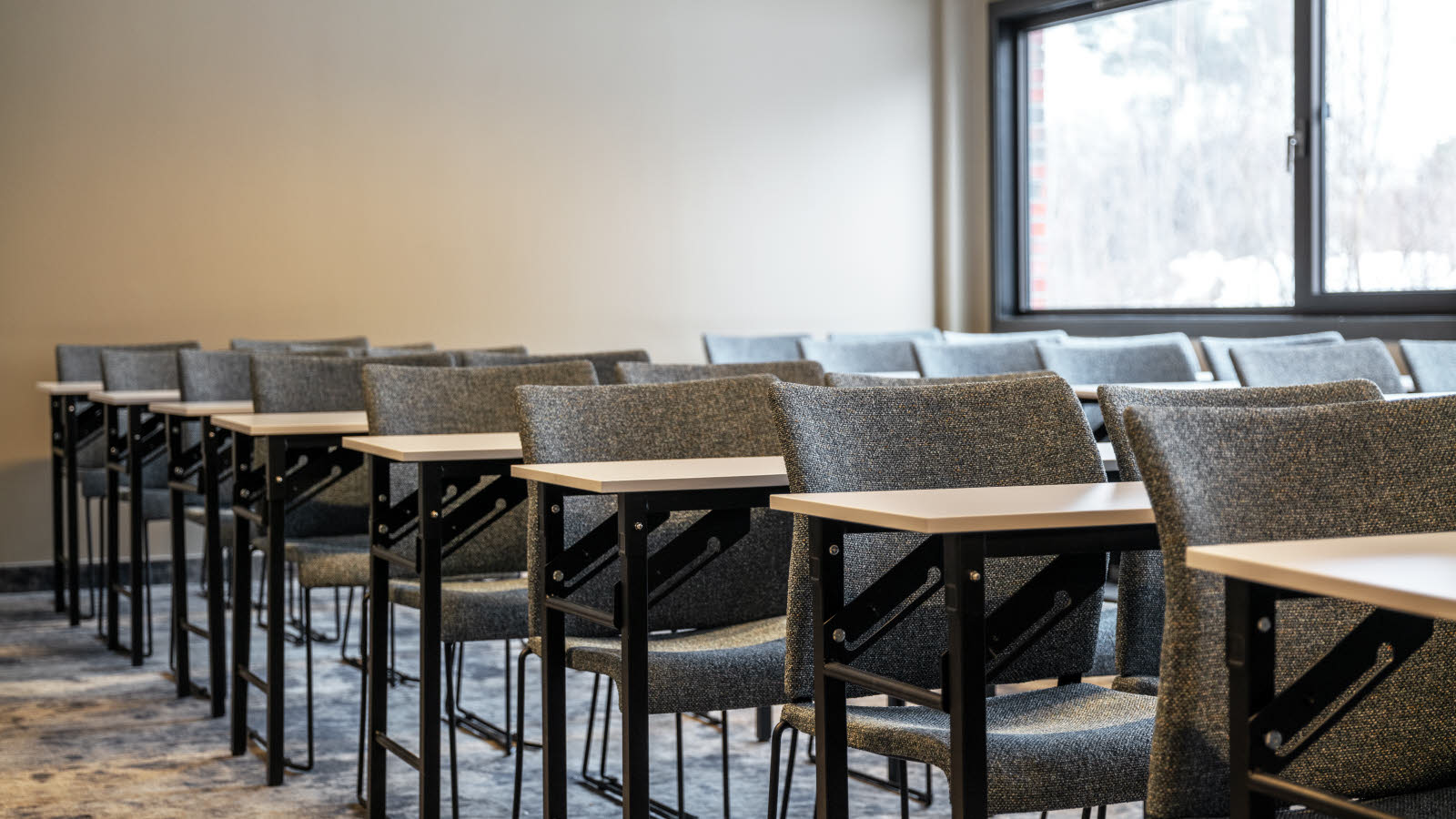 Utsnitt av klasseroms oppsett i møterom. Grå stoler, teppegulv og store vinduer på ene siden.