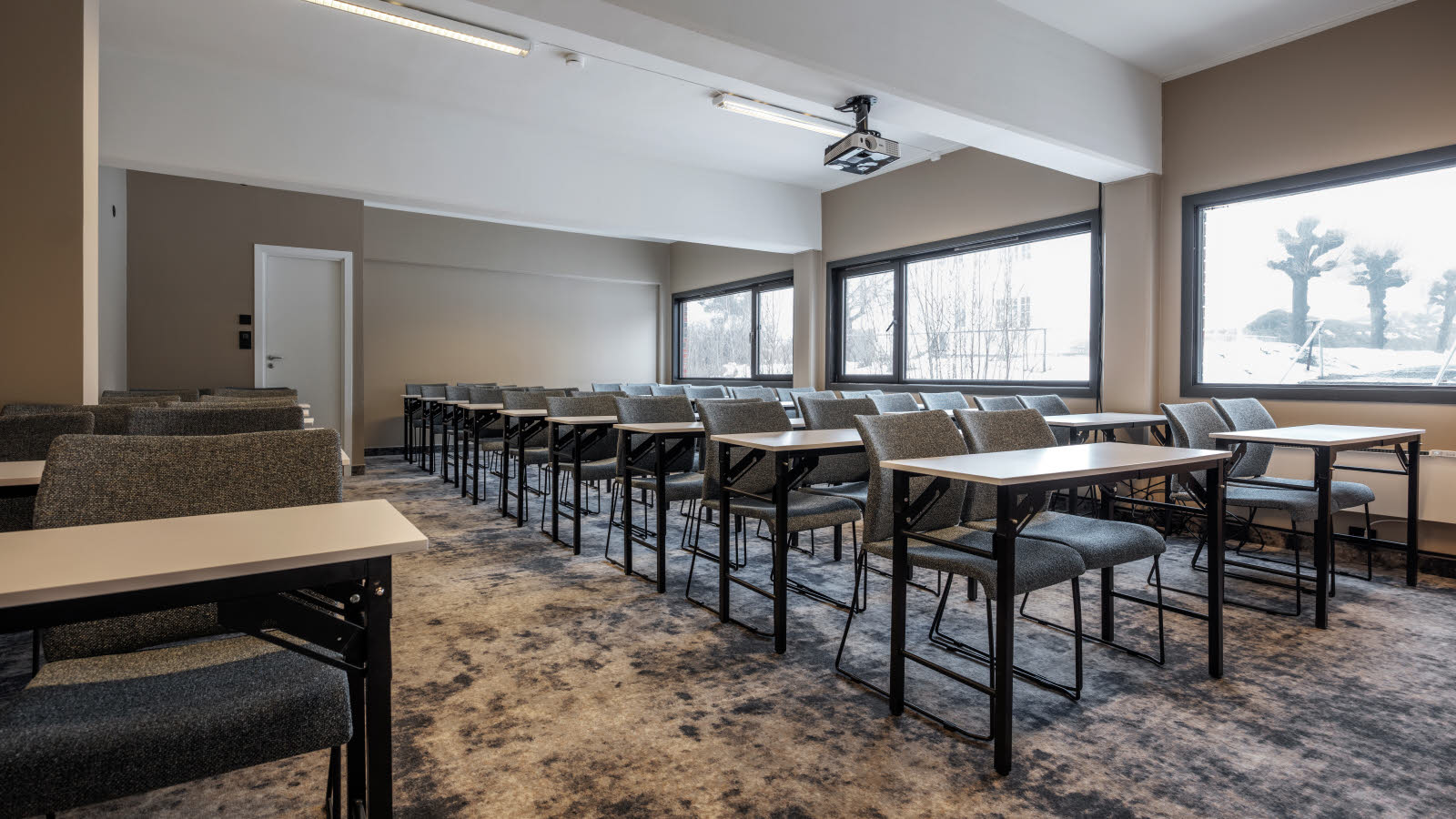 Møterom til 42 personer i klasseromsoppsett, projektor i taket, teppebelagt gulv, utgang bakerst i rommet, vinduer langs langsiden.