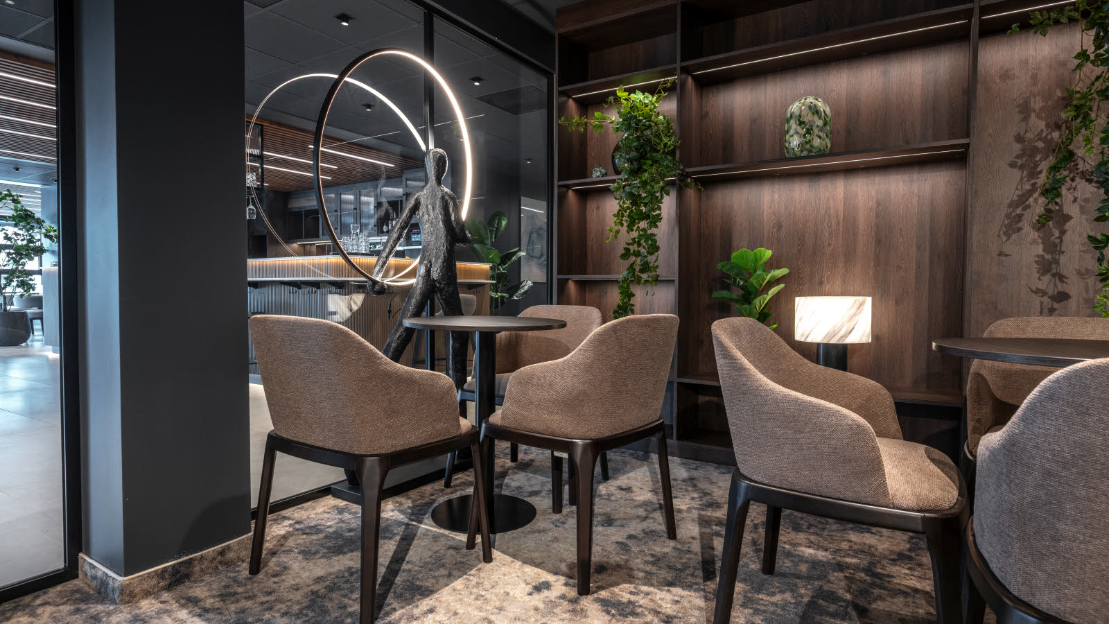 Nærbilde av sitteplasser i et møterom med glassvegg mot lobby. Brun trehylle bakerst med planter og pynt.