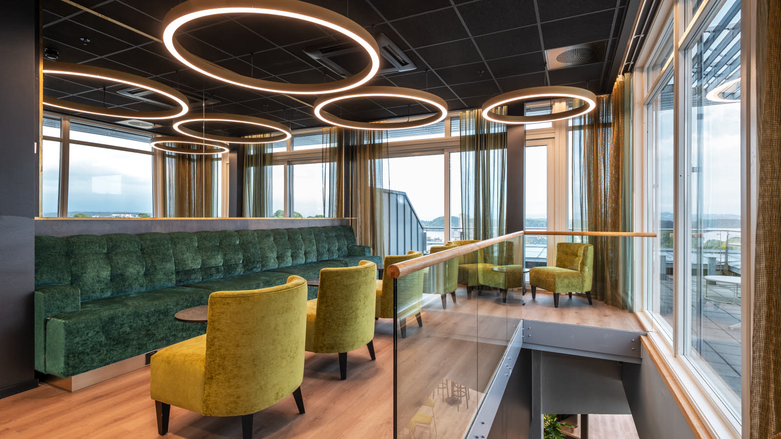Loungeområde i tilknytning et møterom med store grønne lenestoler, grønne sofer og runde taklamper som skaper en god atmosfære. Store vinduer som gir utsikt over byen.