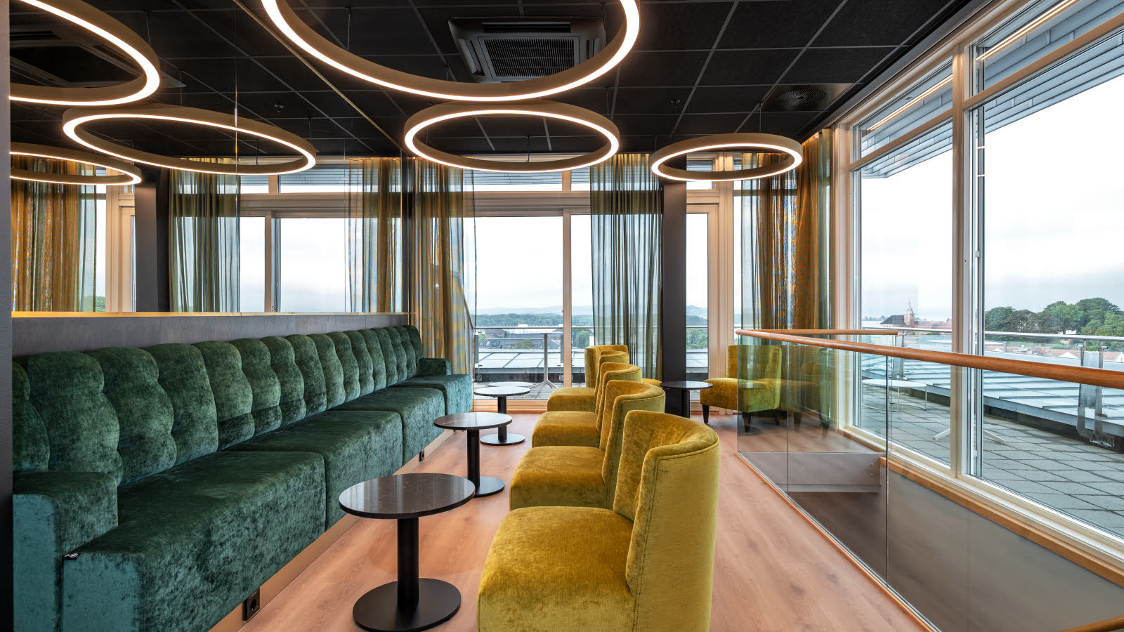 Loungeområde med lang grønn sofa i fløyel og lysegrønne lenestoler, lokalisert i en etasje høyt oppe med store vinduer som gir god utsikt.