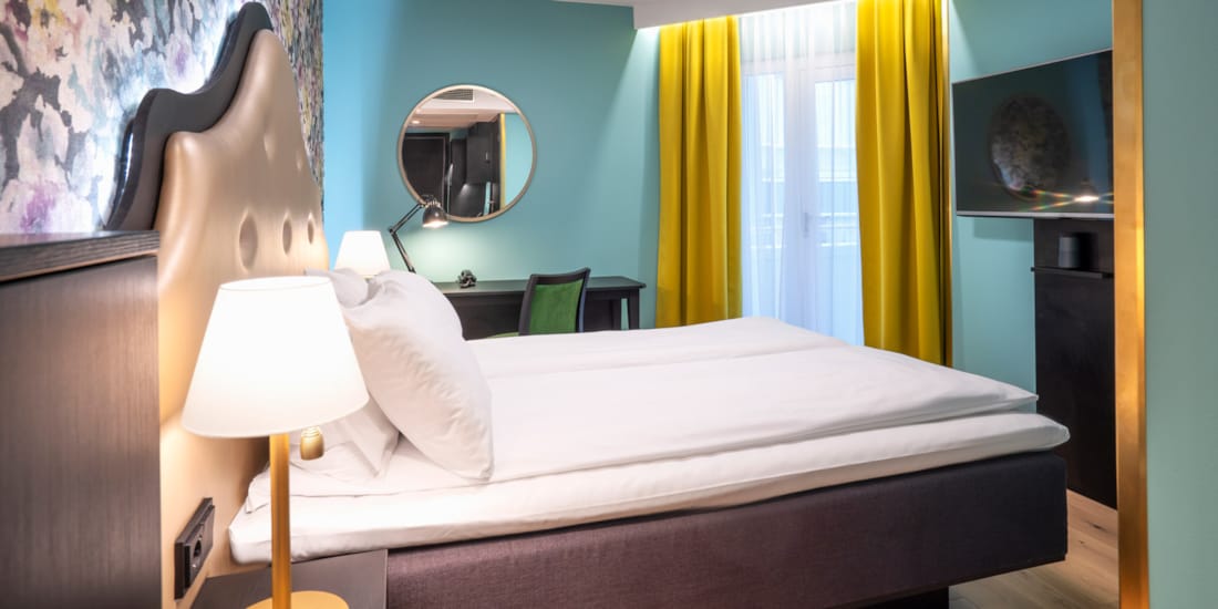 Blomstret tapet bak queen-seng i queen room med turkise farger og gule gardiner, tv og arbeidsplass på Thon Hotel Cecil i Oslo sentrum