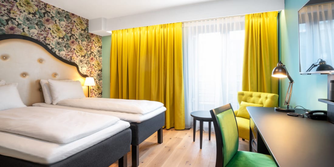 To enkeltsenger, blomstret tapet bak sengene, gul stol, arbeidspult og store vinduer med store gule gardiner på twin room på Thon Hotel Cecil i Oslo sentrum