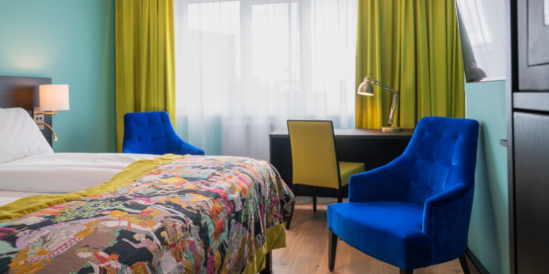 Twin room med hvit sengetøy, mønstret sengeteppe, gule gardiner, skrivepult og blå lenestol på Thon Hotel Europa i Oslo sentrum rett ved Slottsparken