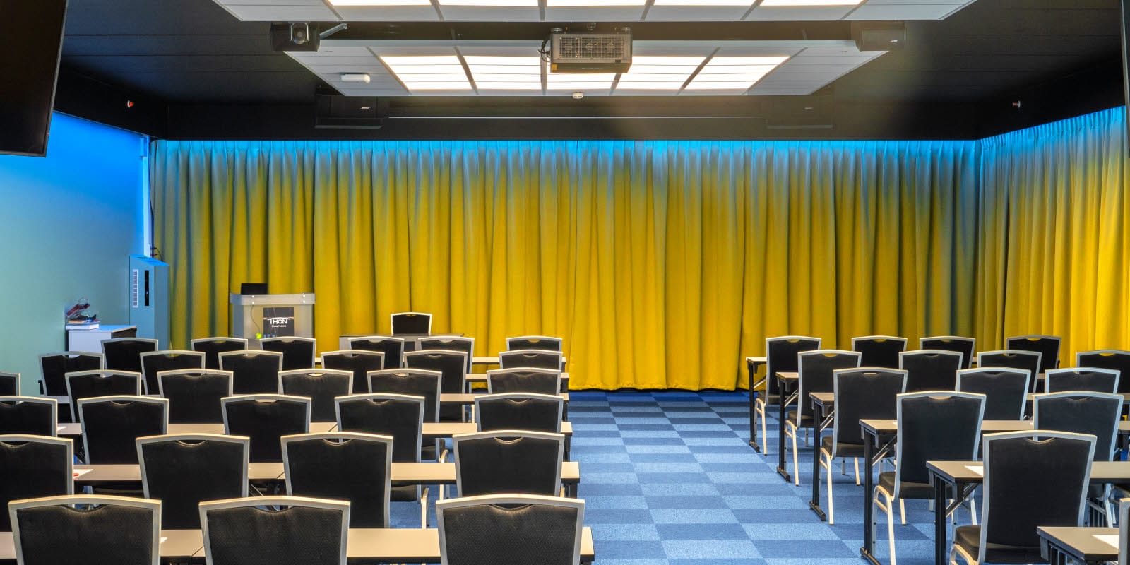 Konferansesal Bjerke satt opp i klasseromsoppsett med plass til 100 stykker. Blå teppe, gule gardiner og prosjektor på Thon Hotel Linne i Oslo