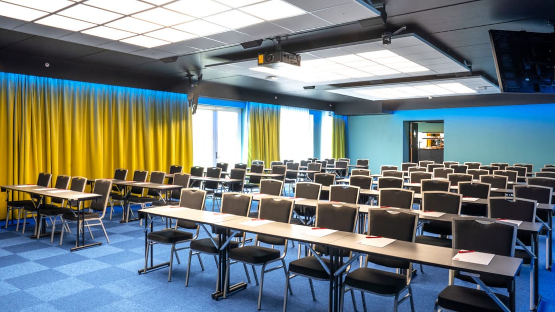 Konferansesal Bjerke satt opp i klasseromsoppsett med plass til 100 stykker. Blå teppe, gule gardiner og prosjektor på Thon Hotel Linne i Oslo