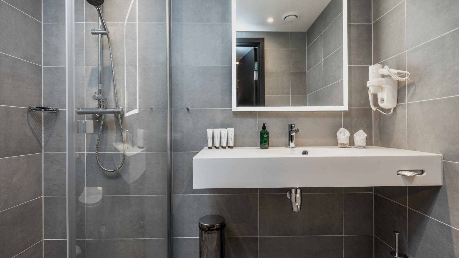 Hotellbaderom med dusj p&#xE5; venstre side og en hvit vask med et stort speil over, gr&#xE5; fliser p&#xE5; veggene. 