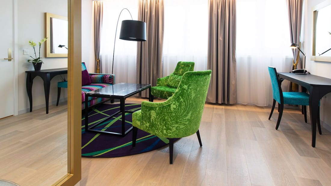 Junior Suite med fargerik teppe, grønne lenestoler, stor stående lampe, speil på veggen og kontorplass