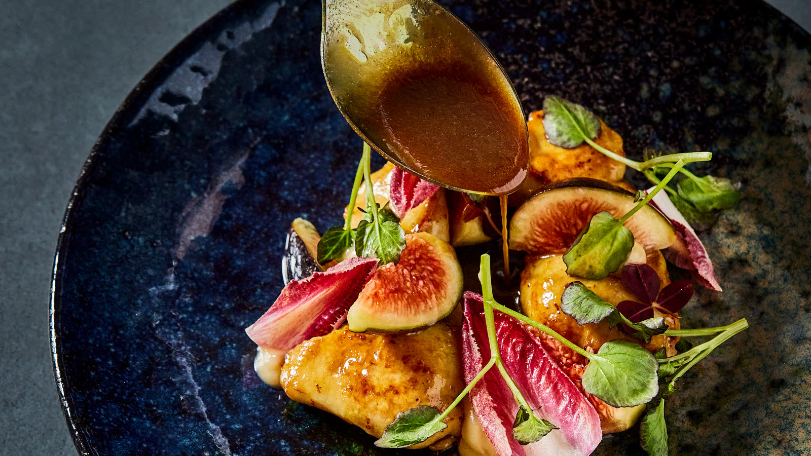 Utsnitt av en tallerken med  fiken, forskjellig salattyper. Spiseskje som heller brun saus over retten.