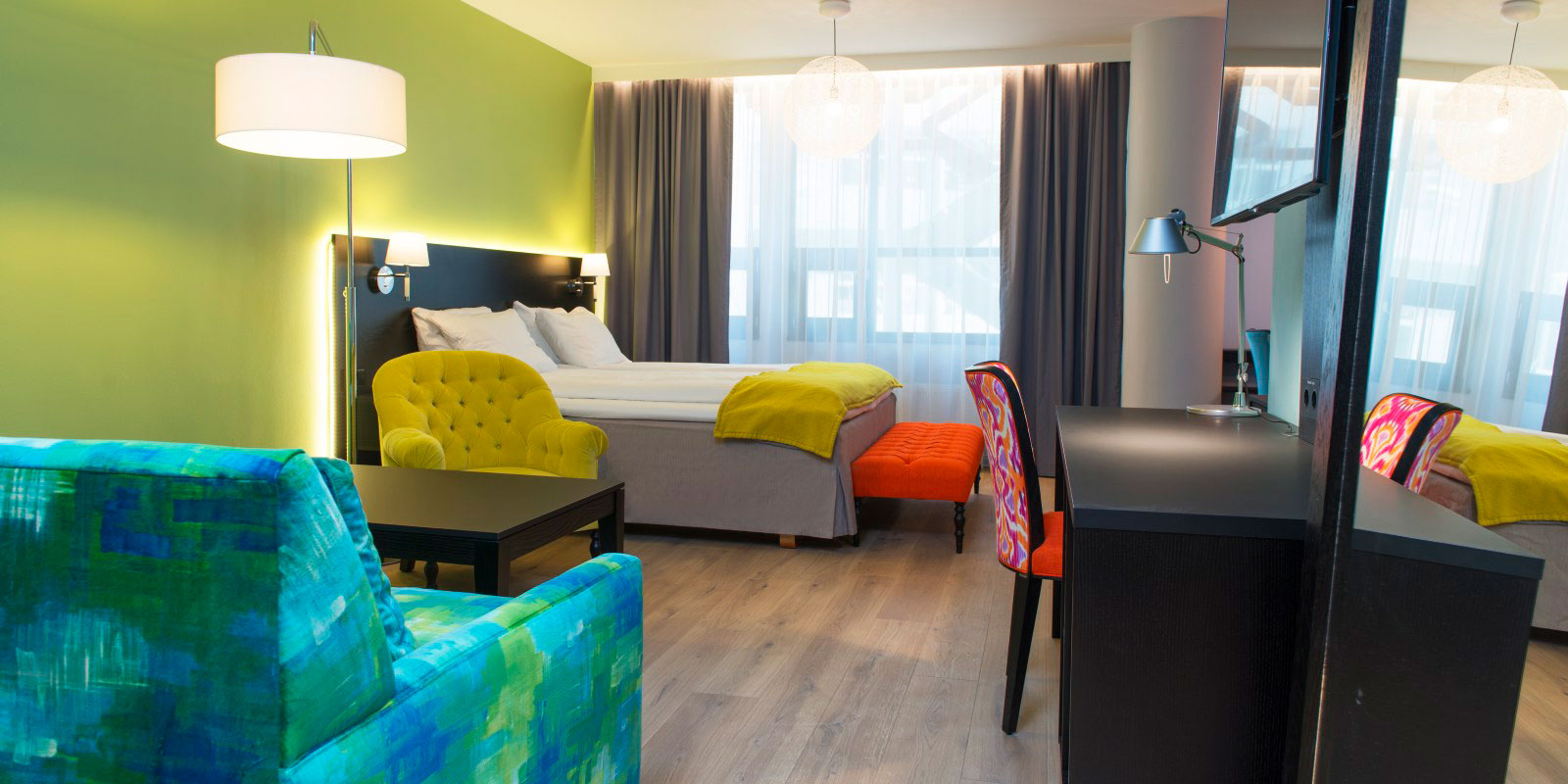 business room dobbeltseng ved vindu, sofa og stålampe og sofabord til ventre, skrivebord me dleselampe, lystette gardiner, parkettgulv